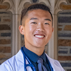 Headshot of student smiling at camera with black hair wearing blue tie, white coat, and stethoscope in front of brick wall