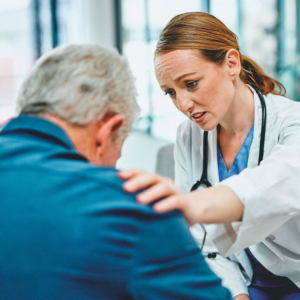 A woman in a white coat comforts an older man, putting a hand on his shoulder. 