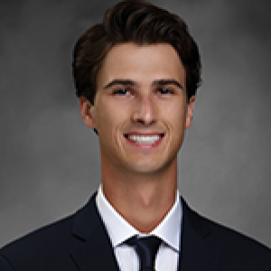 Headshot of Casey Gilles smiling at camera in front of grey backdrop wearing black suit and navy blue tie
