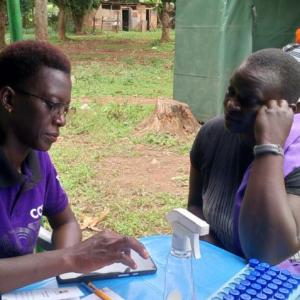 Two women sit at a table outdoors facing each other, one using a mobile phone.