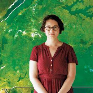 A woman in a burgundy dress stands in front of a green map.