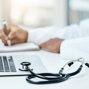 A person in a white coat sits at a laptop with a stethoscope nearby