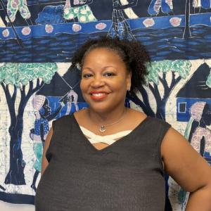 A Black woman stands in from of a blue, green, and black mural, smiling.