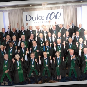 Portrait of elder alumni in green regalia with a Duke 100 centennial banner in the background