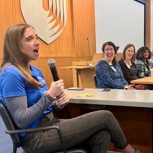 a student in the foreground speaking on a mic with faculty in the background laughing