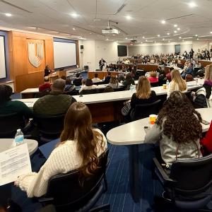a lecture hall full of guests looking on as a staff member talks to them at the podium