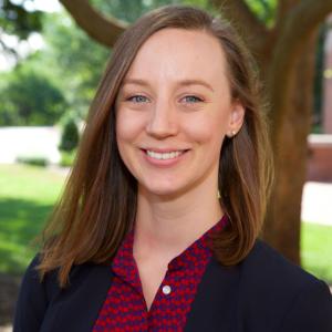 Headshot of Carolyn Gilbert smiling at camera with should length mousy-brown hair, wearing red printed shirt under navy blazer, standing in front of a tree