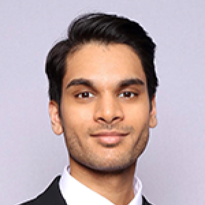 Headshot of Sunag Udupa wearing black suit and black tie slightly smiling at camera in front of grey backdrop