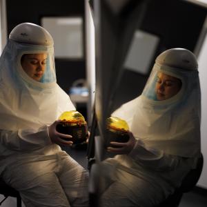 A researcher in full-coverage protective garb handles a sample beside a shiny surface that reflects her image.