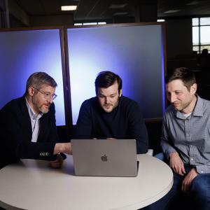Duke researchers Jonathan Posner, Elliot Hill, and Matthew Engelhard sit at a small round table looking at a laptop screen