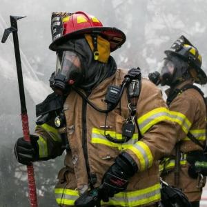 Two firefighters in heavy gear and gas masks fight a fire.