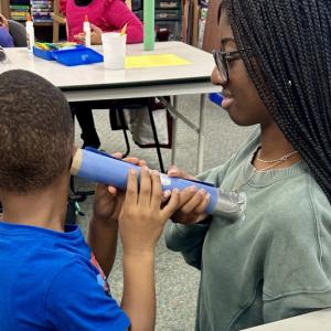 An elementary school student uses a homemade stethoscope to listen to a medical student's heart.
