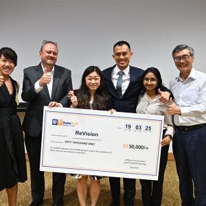 Image: Six people holding a large ceremonial check for $50,000 made out to "ReVision." They are standing in front of a projector screen, smiling and giving thumbs up.