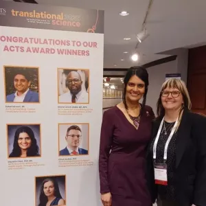 Gayathri Devi standing with ACTS Executive Director Kim Stelmaszak beside a large poster showing this year's ACTS Award winners