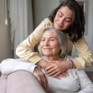 Older mother seated with daughter above with her arms around her mother