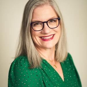 Professional headshot of a person with shoulder-length light hair, dark-framed glasses, and a green blouse.